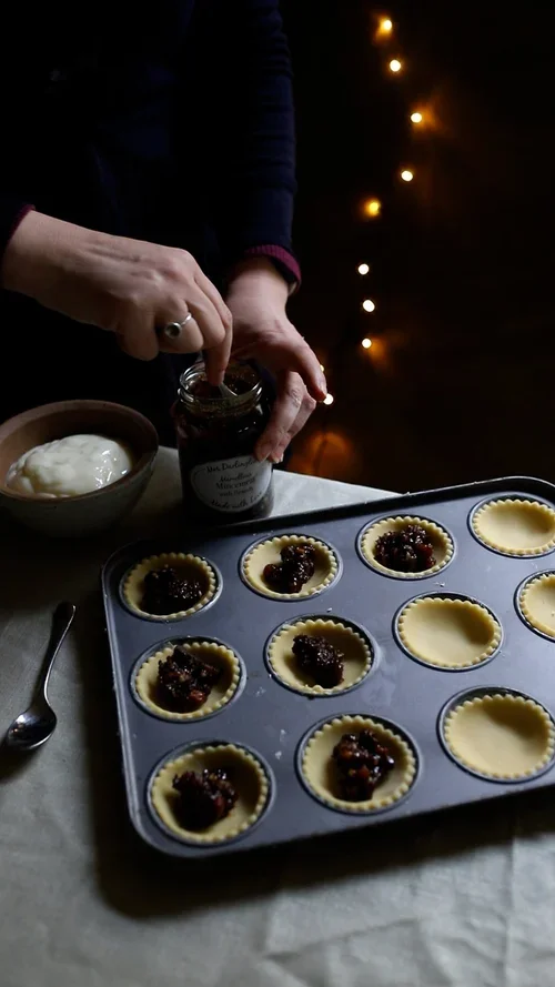 Person scooping dark fruity mincemeat from a glass jar into raw pastry cases in a metal baking tin on a linen tablecloth