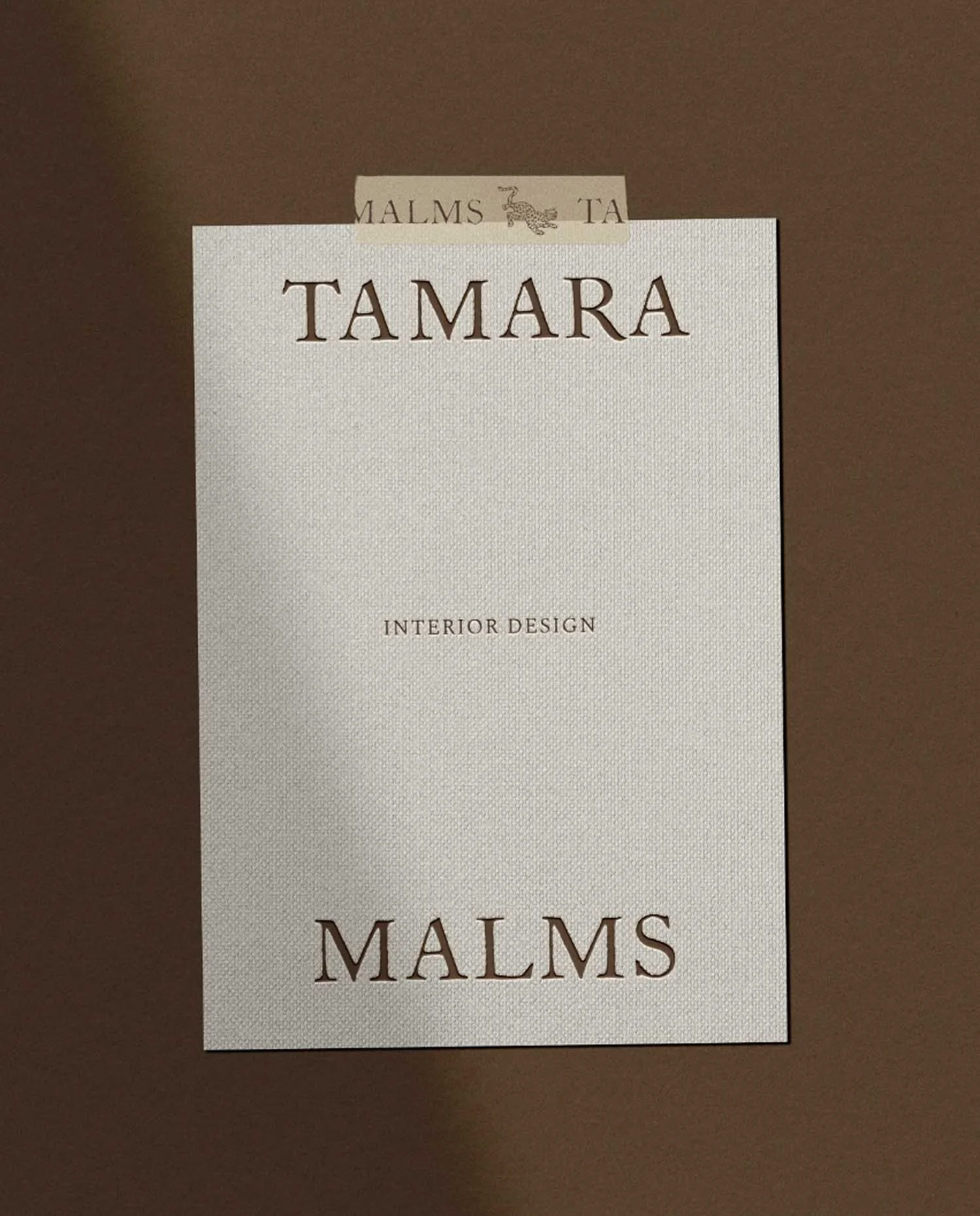 A cream textured card with the words Tamara Interior Design Malms held against a dark brown wall with branded washi tape