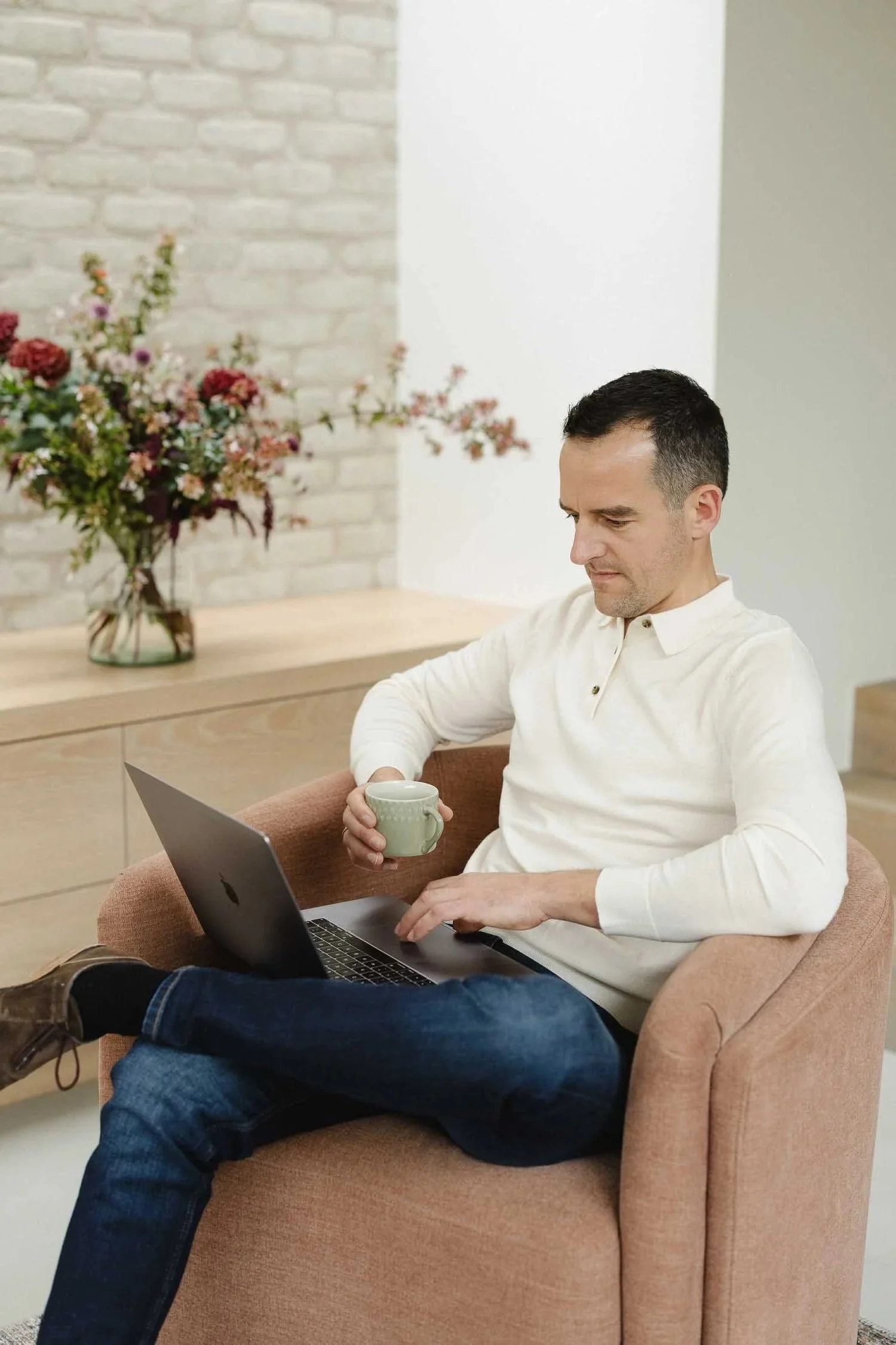 A man in a white polo shirt sits in a pink armchair holding a green cup while working on a laptop near a vase of flowers