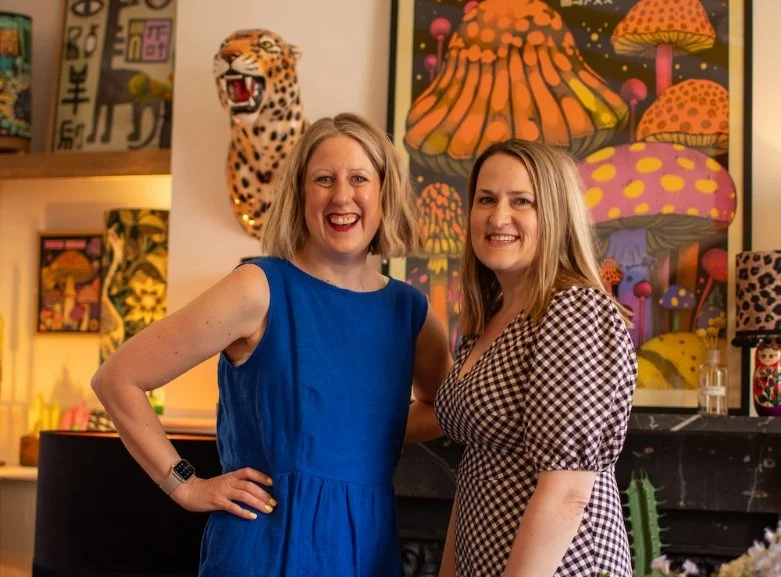 Two women smiling in front of a leopard head mount and vibrant mushroom artwork in a room with eclectic and bold interior