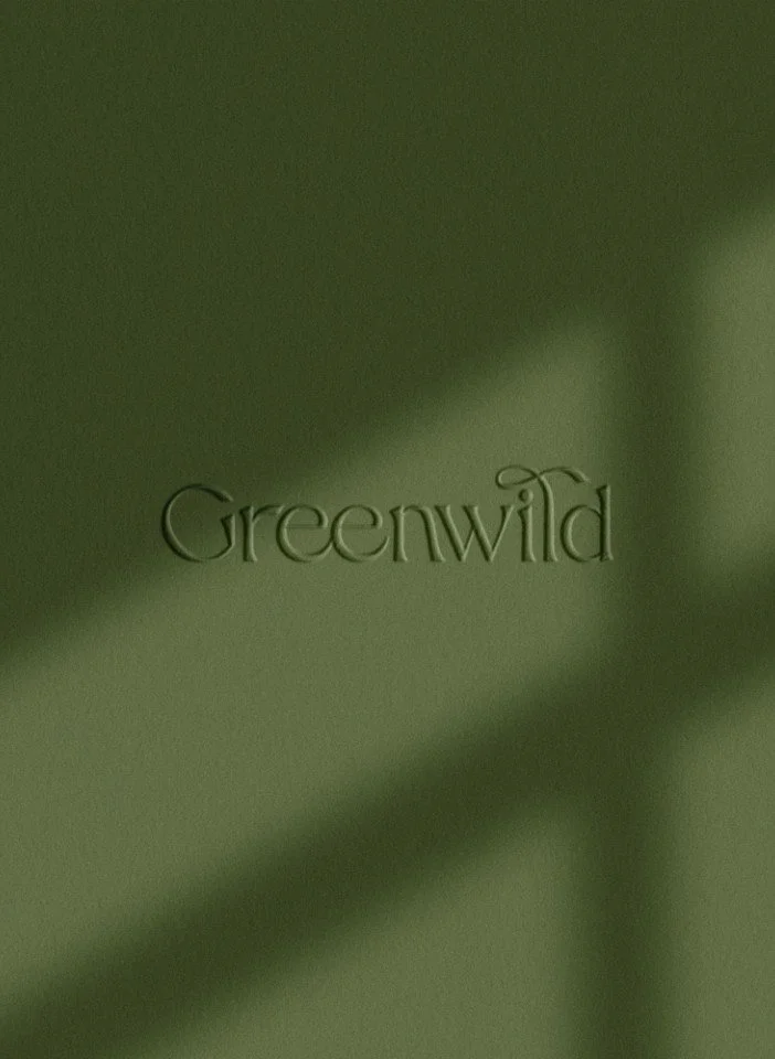 Close up of an olive green textured surface with the Greenwild logo debossed in the centre under soft leaf shadows