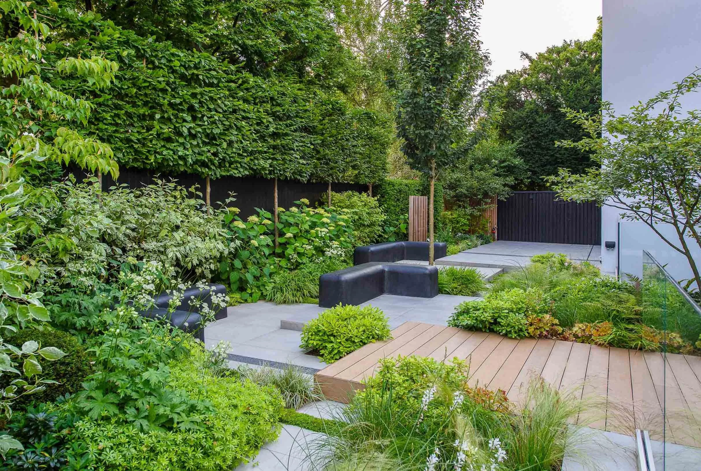 Modern garden featuring grey stone paving black minimalist benches a timber deck and lush green trees behind a dark fence