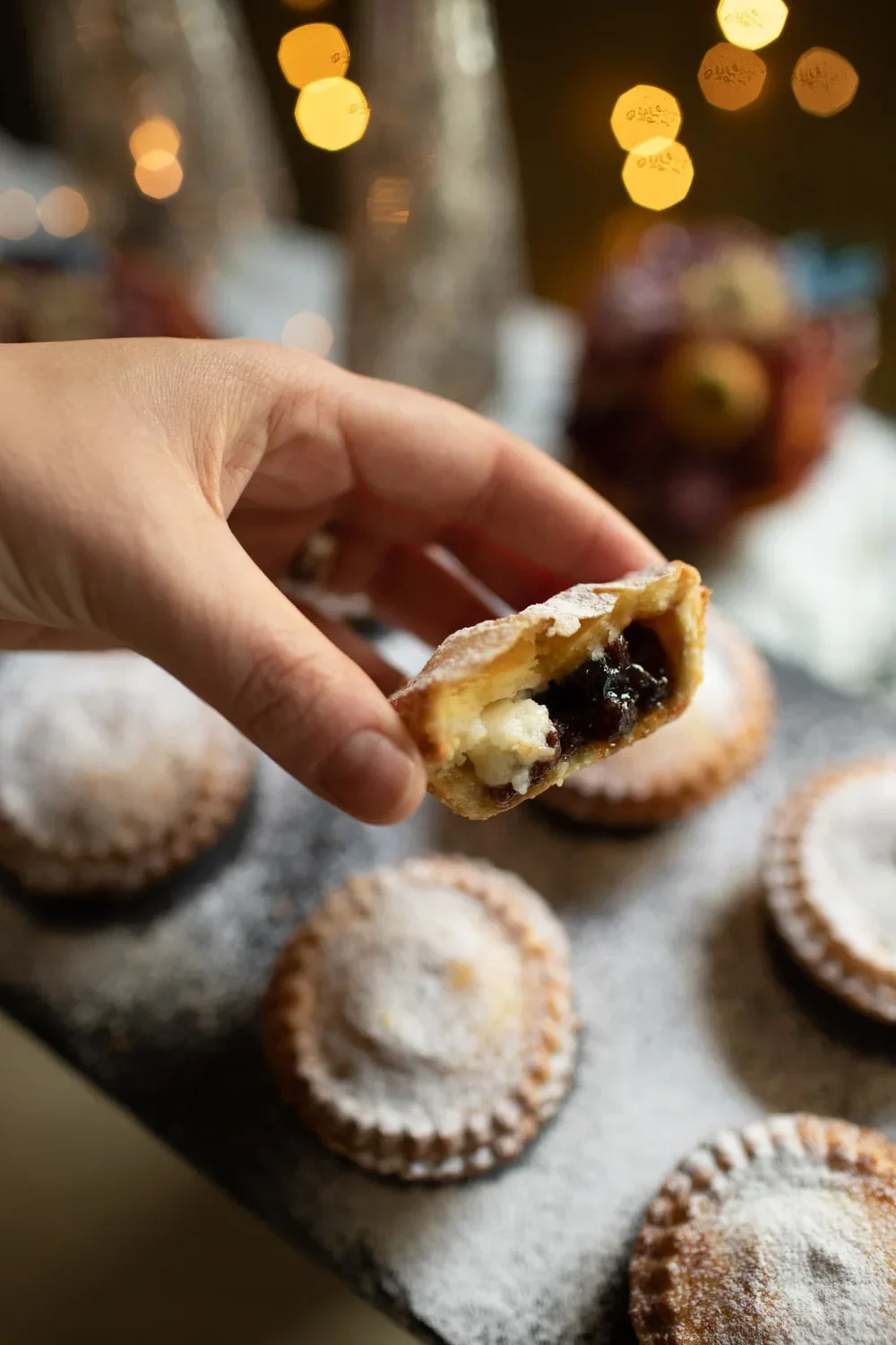 A hand holding half a mince pie showing the dark fruit and white cream cheese filling against a festive bokeh background