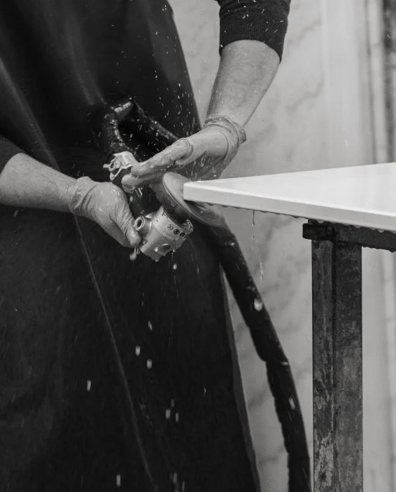 A craftsman in protective gear using a hand tool with water to polish the edge of a white stone slab in a workshop