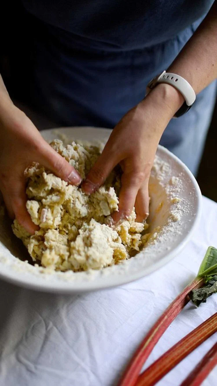 Close-up of hands mixing a shaggy scone dough in a white ceramic bowl with fresh pink rhubarb stalks on the side