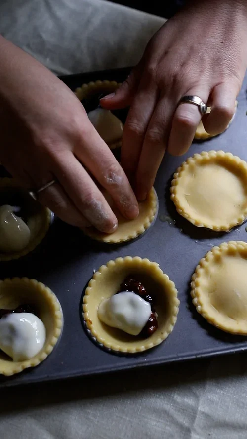 Close up of hands pressing a fluted pastry lid onto a mince pie base filled with dark mincemeat and white cream cheese sauce