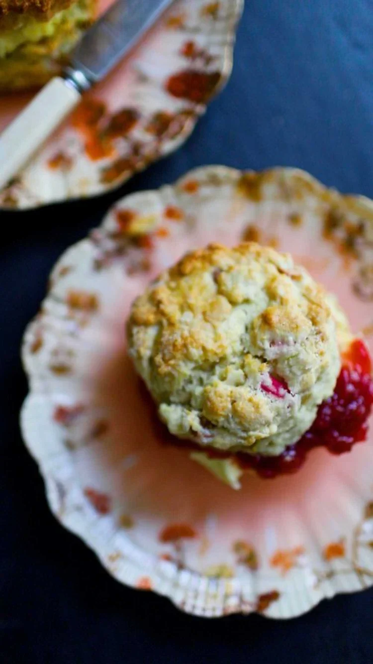 Close-up of a fruit scone on a decorative pink plate with a generous layer of red jam and a vintage bone-handle knife