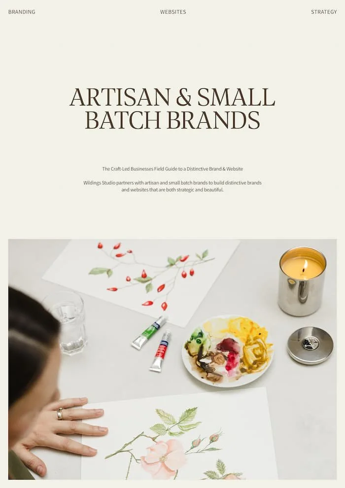 Artist painting botanical illustrations of roses and berries on a white desk with paint tubes a palette and lit candle