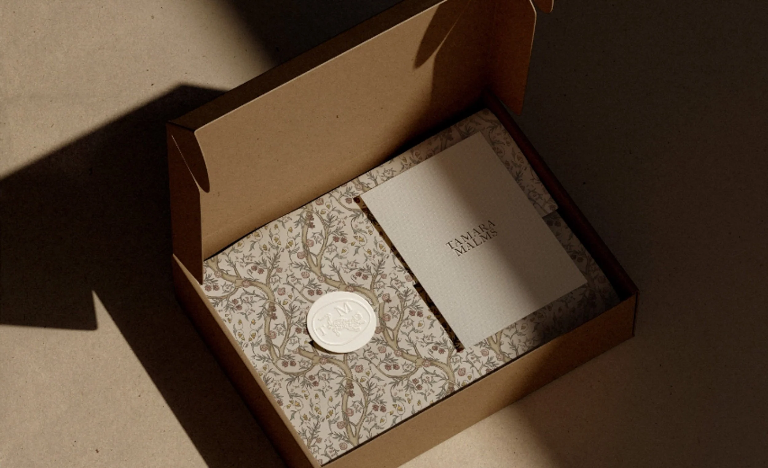 Open cardboard mailer box containing patterned tissue paper a white wax seal and a Tamara Malms branded information card