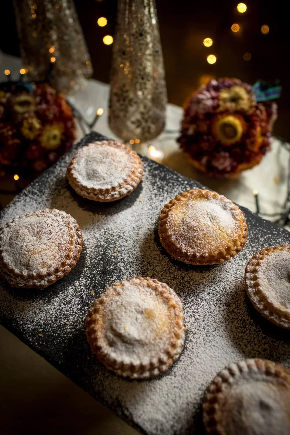 Five golden mince pies with fluted edges dusted in icing sugar on a slate board with festive decorations in background