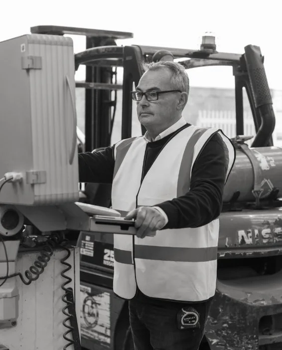 Black and white photo of a worker in a high visibility vest and glasses operating a control panel in a stone warehouse