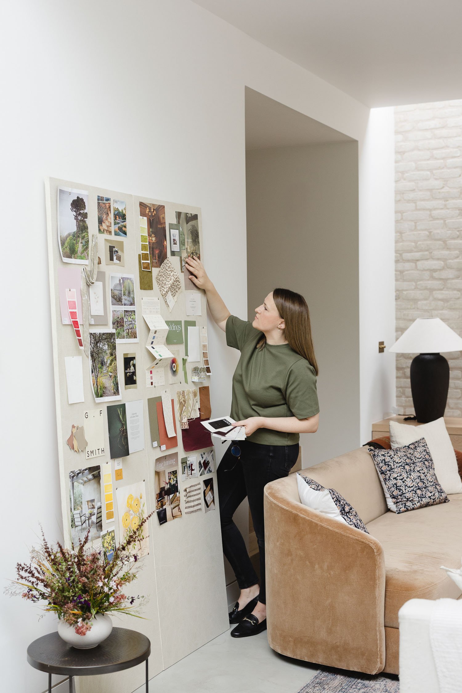 A woman in an olive green shirt pins various fabric swatches and architectural photos to a large cream pinboard in a room