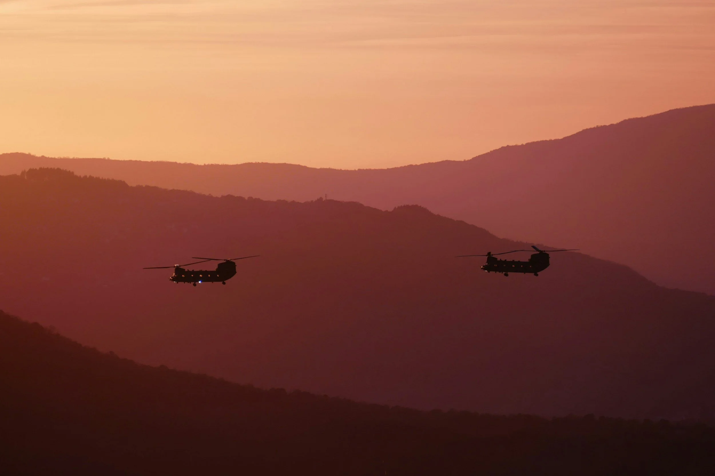 Two helicopters flying over a mountainous landscape at sunset or sunrise.