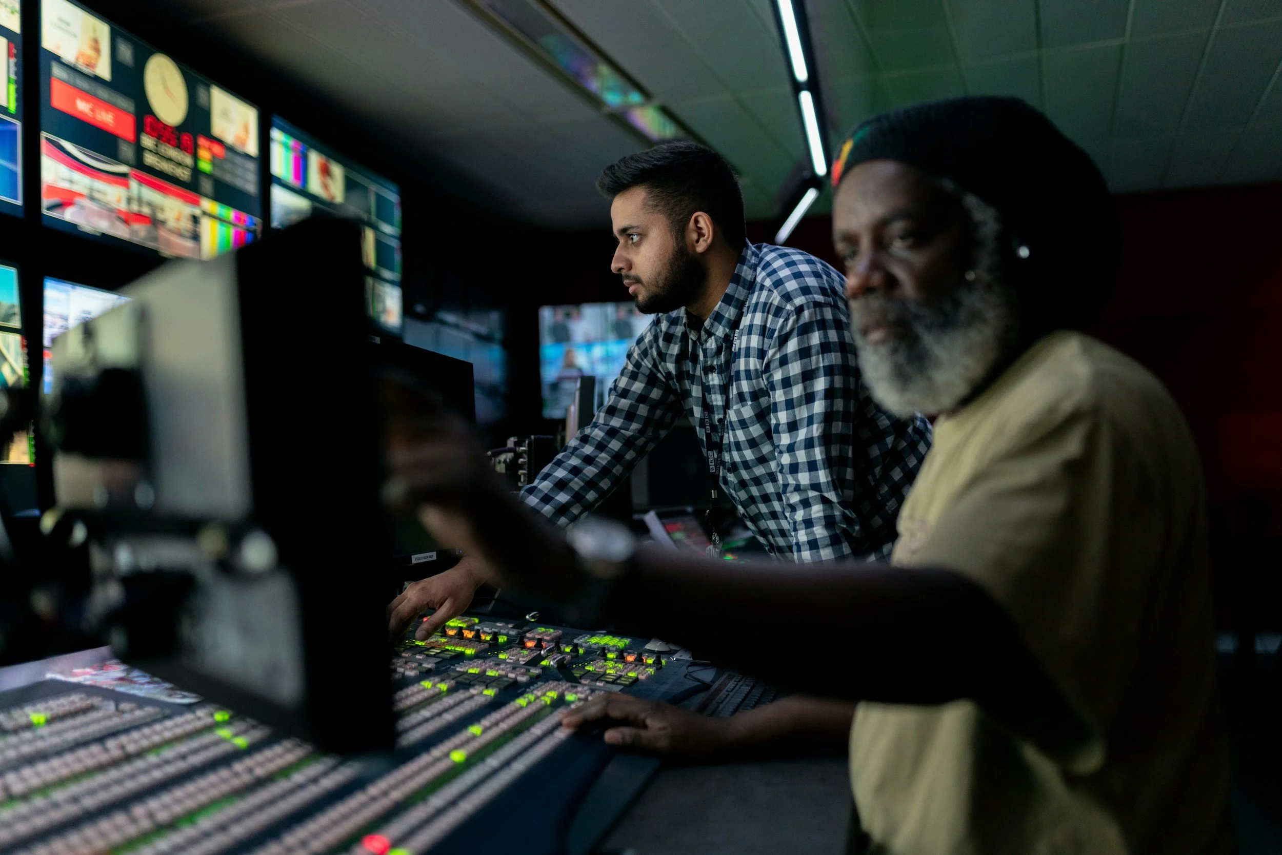 Two men working at a broadcast control console with multiple monitors in a darkened room.