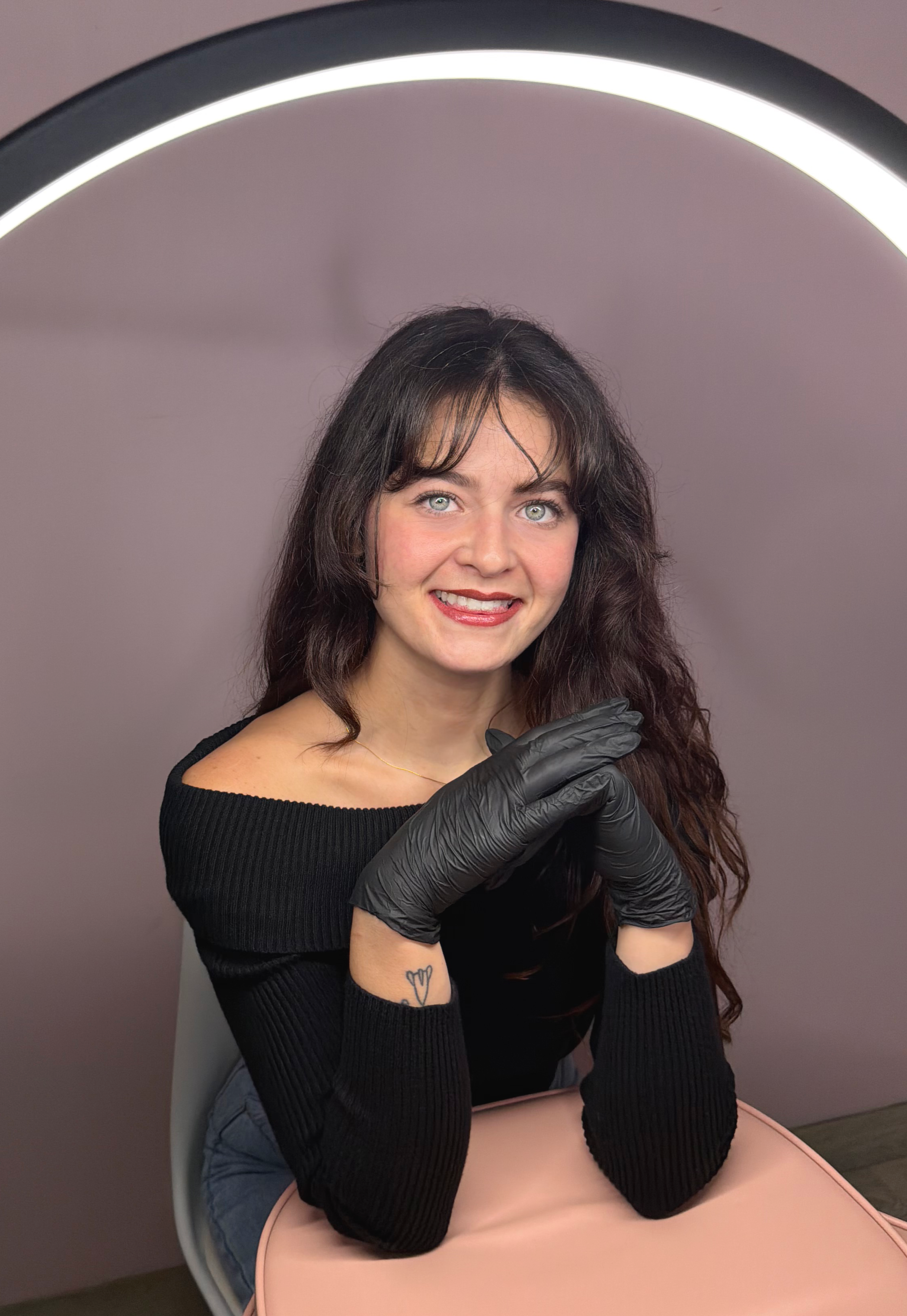 A woman with long dark hair, blue eyes, and a black off-shoulder top, wearing black gloves, sitting at a pink table, smiling at the camera, with a ring light overhead.