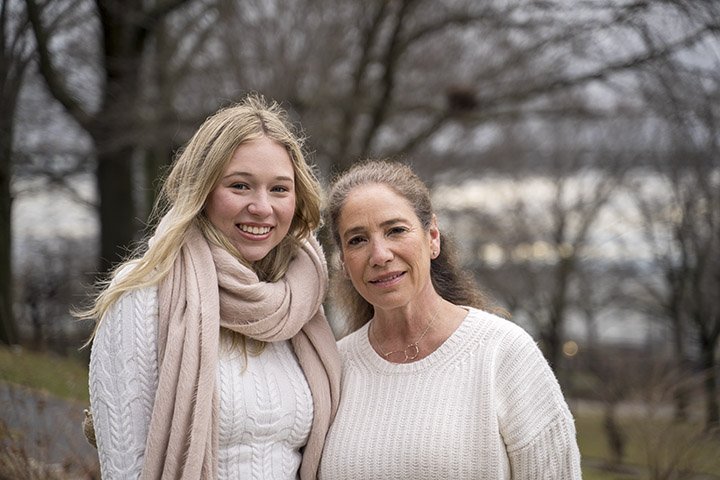 A young woman and an older woman standing outdoors in a park with bare trees, smiling at the camera.