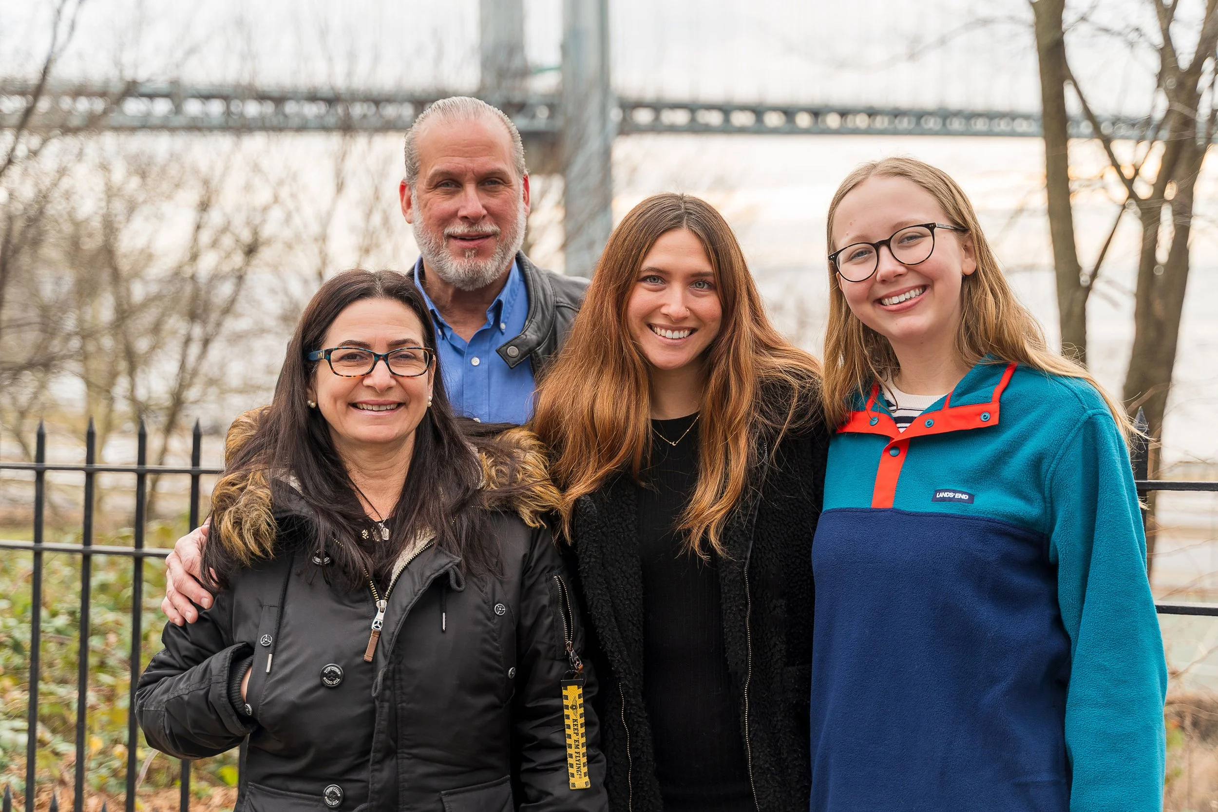 Group of five people smiling outdoors behind a black metal fence with trees and a bridge in the background.