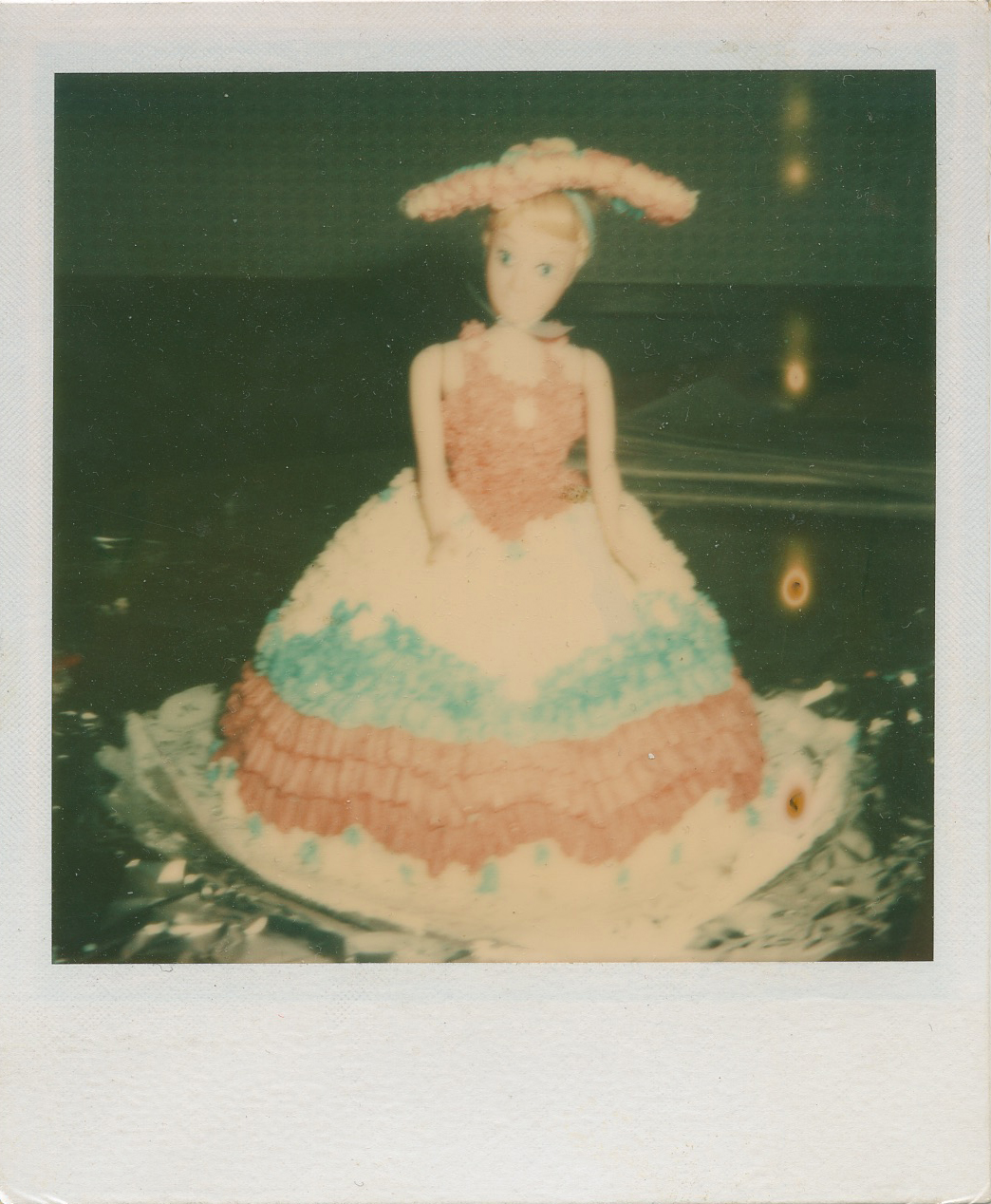 Vintage photo of a doll dressed in a pink, white, and blue ball gown with a matching pink hat, displayed on a reflective surface.
