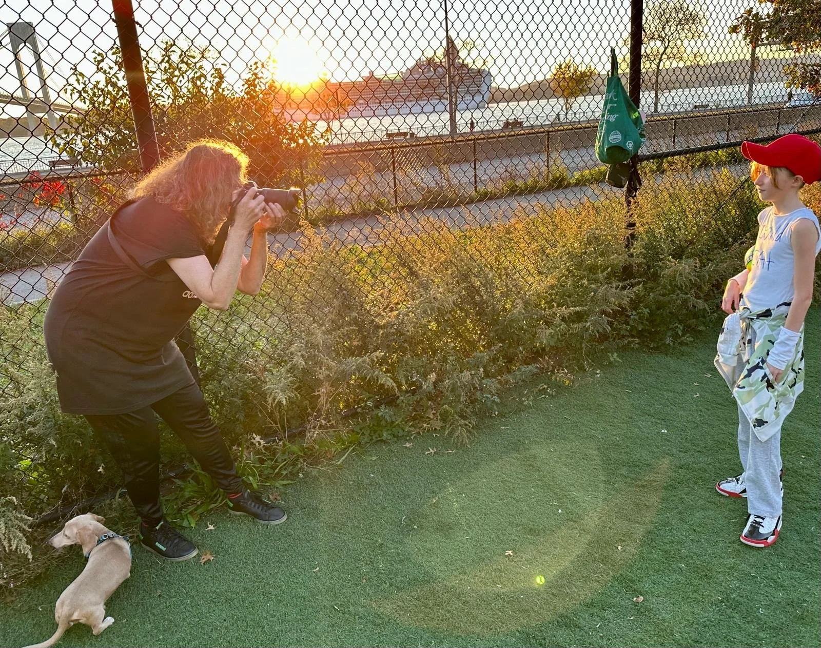 A woman taking a photograph of a young boy wearing a red cap near a chain-link fence during sunset, with a small dog sitting on the ground nearby, and a tennis ball on the grass.