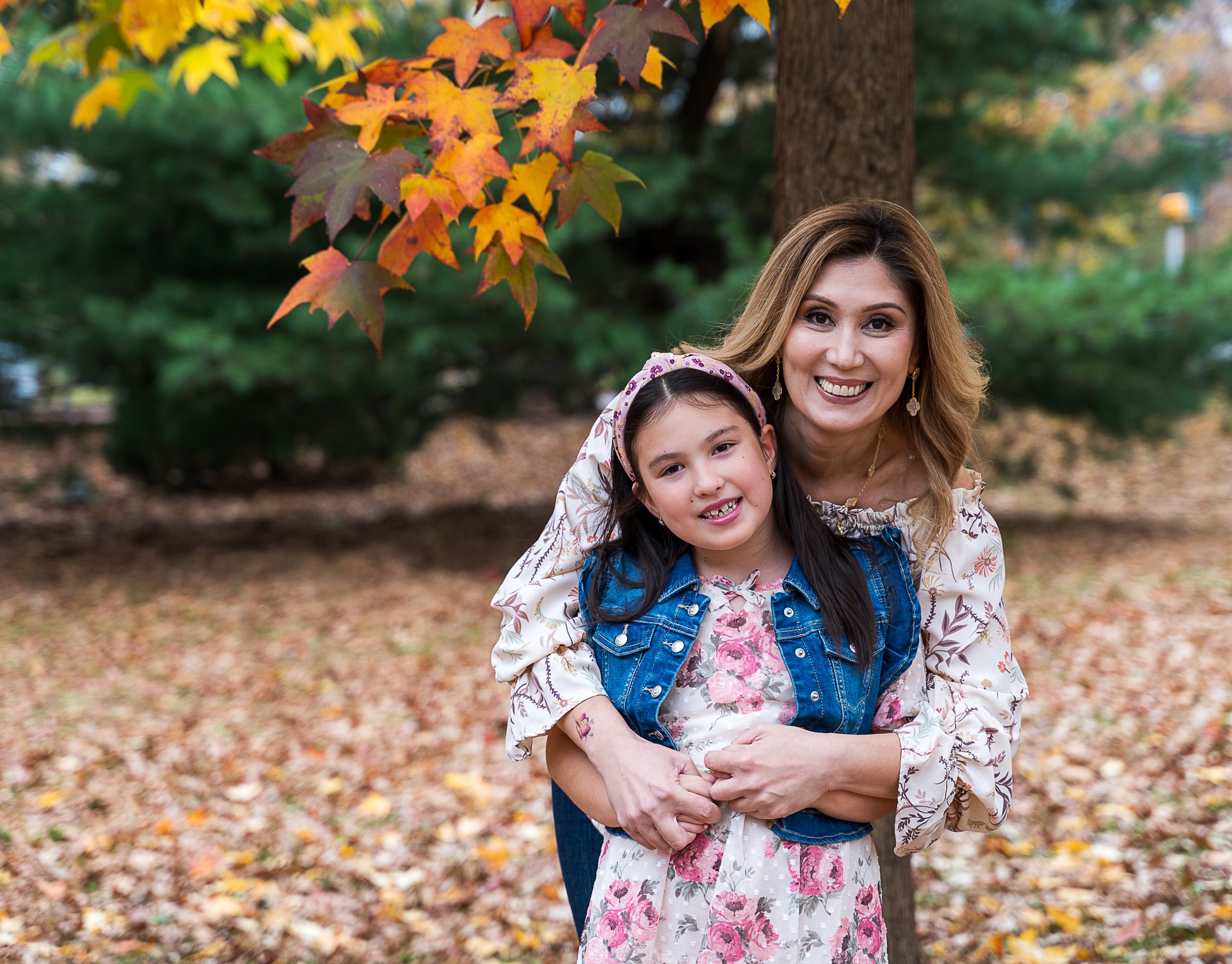 A woman and a young girl standing outdoors among falling autumn leaves, smiling at the camera. The woman has blonde hair and is wearing a floral top, while the girl has dark hair, a pink headband, and is dressed in a floral dress with a denim vest.