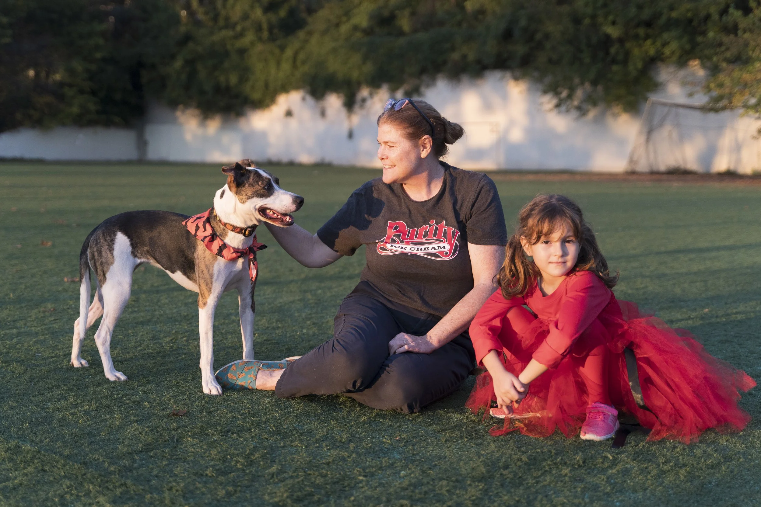 A woman, a girl in a red dress, and a dog are sitting on a grassy field during sunset. The woman is petting the dog, and the girl is looking at the camera.