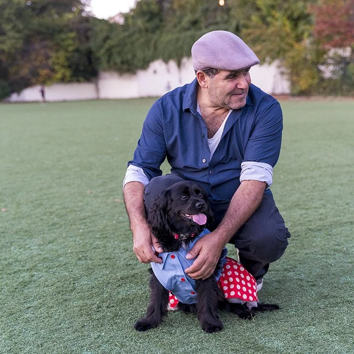 A man wearing a purple cap and a blue shirt crouches on the grass, holding a black dog dressed in a polka dot outfit with red and white colors. The man is outdoors with trees and a white fence in the background.