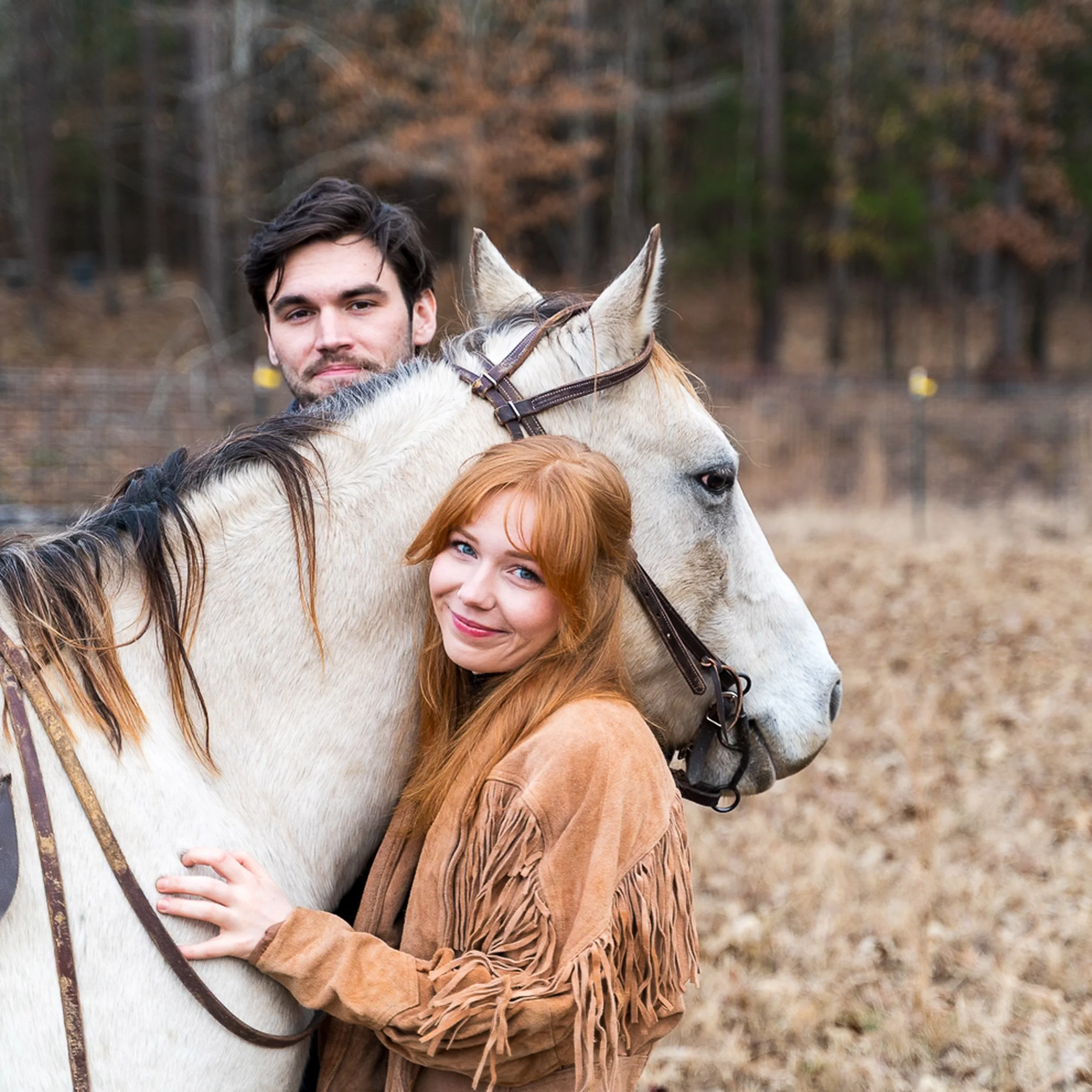 A young man and woman hugging a white horse outdoors in a wooded area with fall foliage.