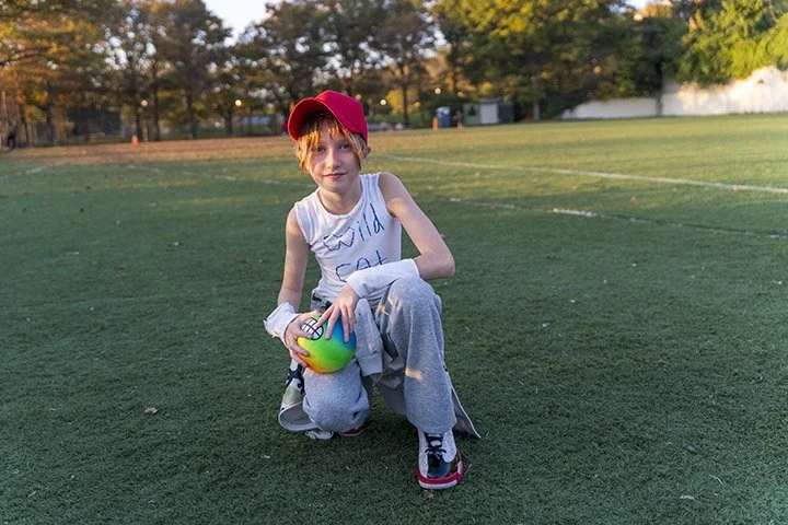 A young boy crouching on a sports field holding a multicolored ball, wearing a red cap, white sleeveless shirt, gray pants, and sneakers.