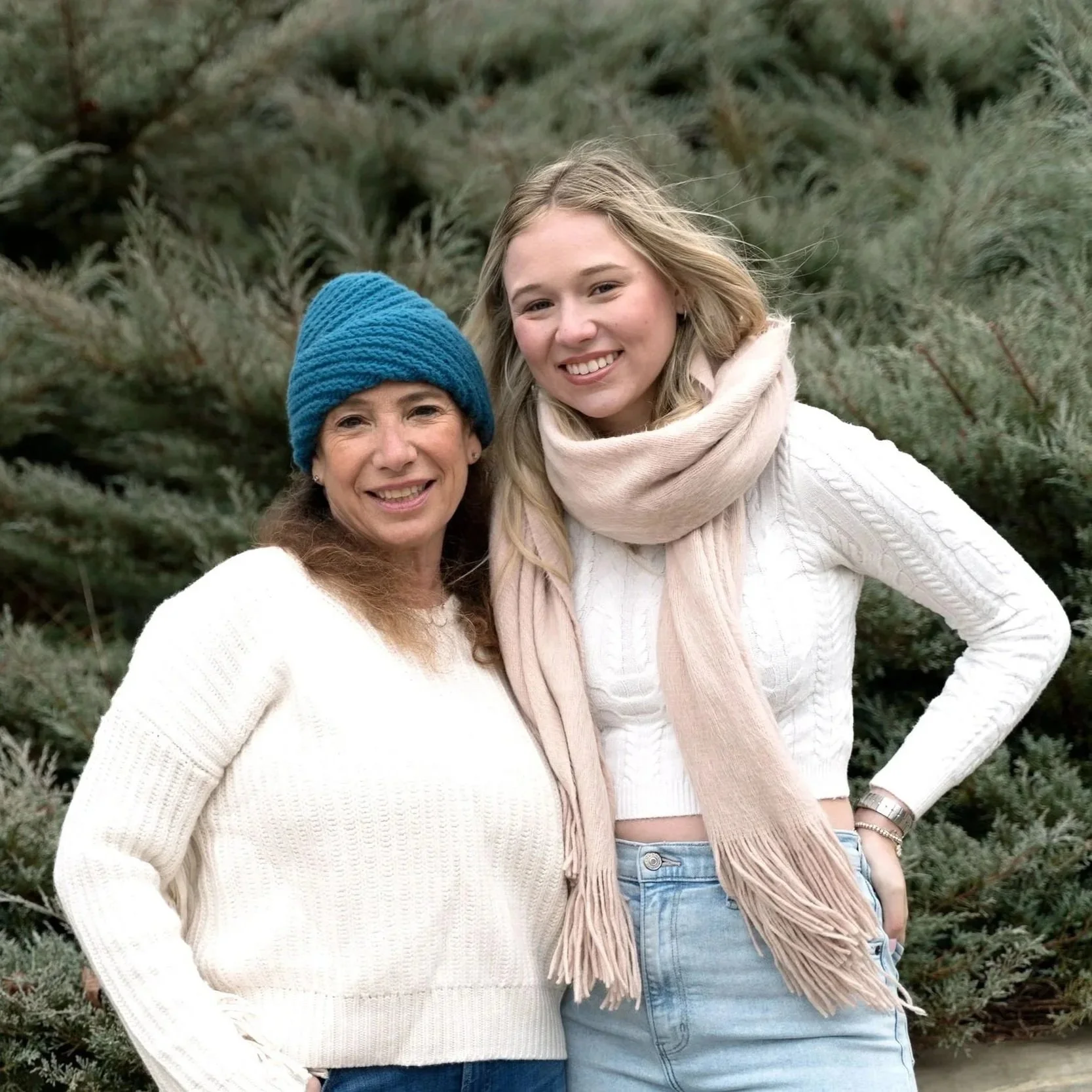 Two women standing outdoors among green shrubs, smiling at the camera. One woman is wearing a blue knit hat and cream sweater, and the other is wearing a beige scarf and cream knit sweater.
