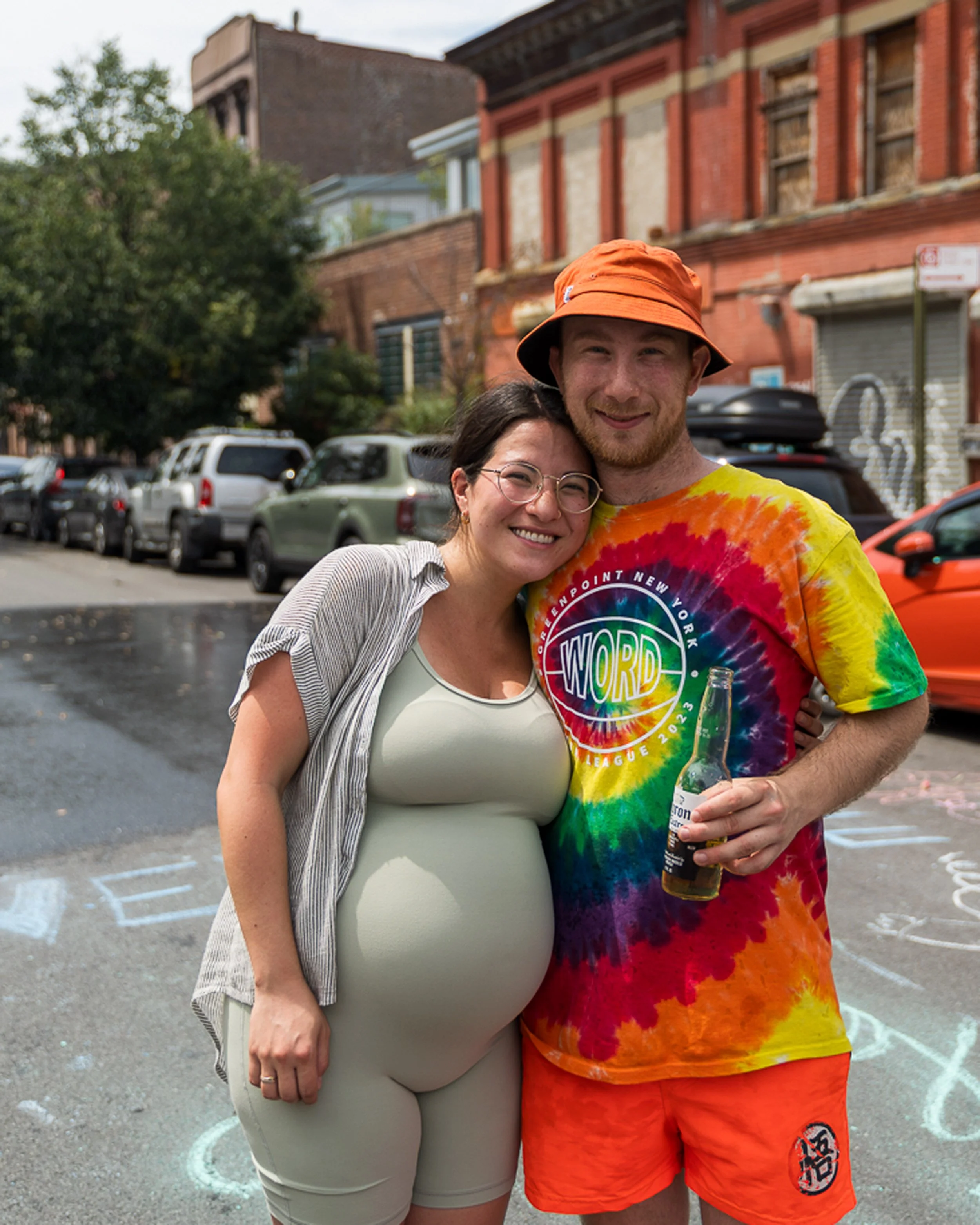 Two smiling people, a woman and a man, hugging outdoors on a city street, with parked cars and buildings in the background. The woman has short dark hair, glasses, and is wearing a light-colored outfit. The man has red hair, a beard, and is wearing a multicolored tie-dye shirt with a logo, orange shorts, and an orange hat. The man is holding a bottle of beer.