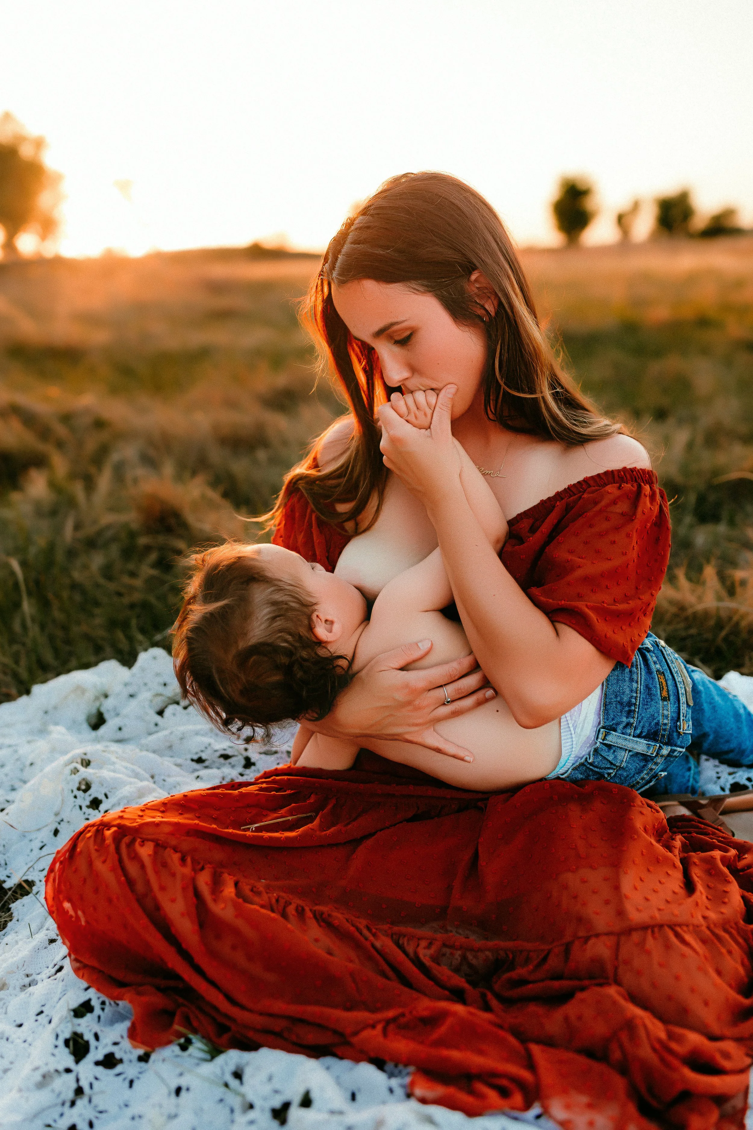 A woman with long brown hair wearing a red dress is sitting on a blanket outdoors during sunset, breastfeeding a young child with curly brown hair and no shirt, in a grassy field with scattered trees in the background.