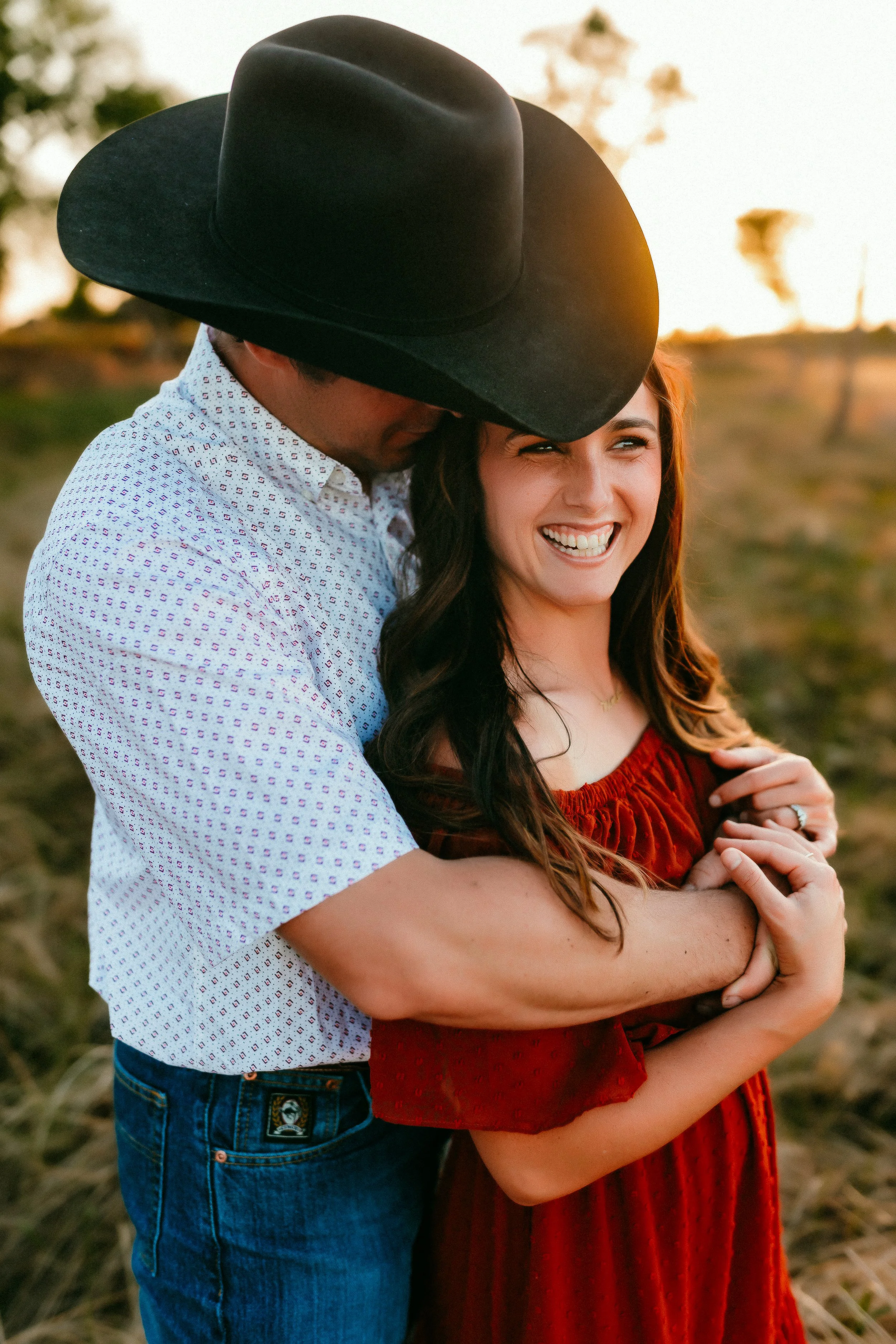 Couple embracing outdoors at sunset, woman smiling and man wearing a black cowboy hat, in a rural setting.