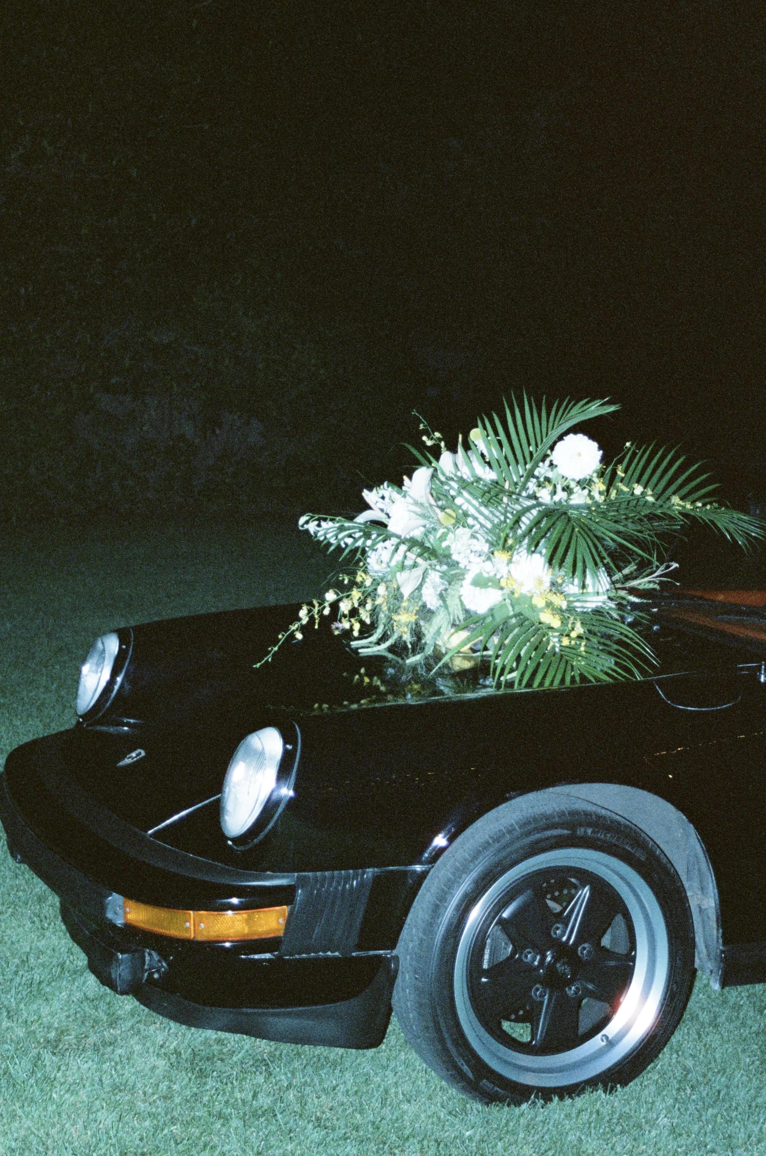 A black vintage Porsche car with a large bouquet of white and yellow flowers on the hood, parked on grass at night.