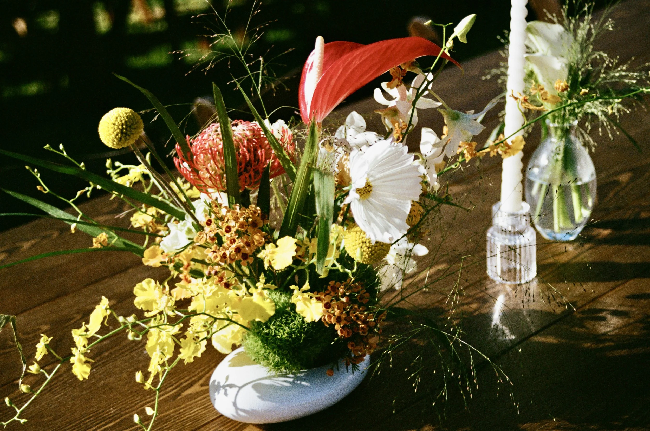 Colorful flower arrangement in a white vase on a wooden table, with a glass vase holding more flowers nearby.