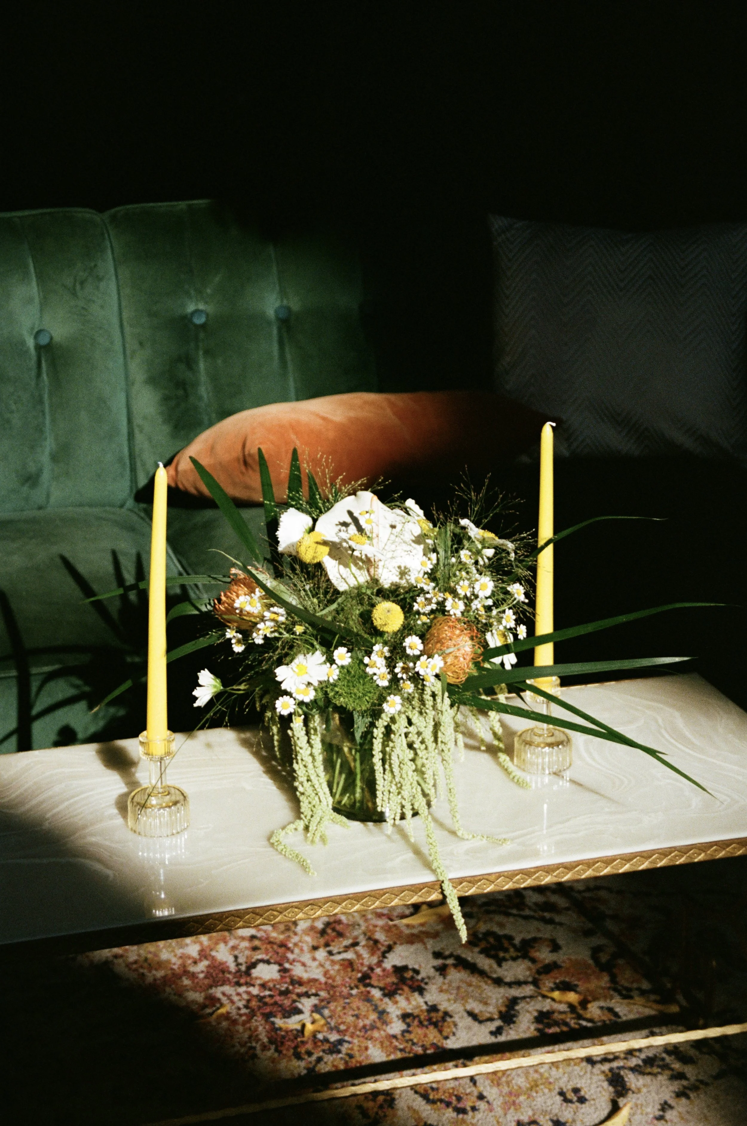 A floral arrangement with yellow and white flowers, two yellow candles in glass holders, on a white tablecloth in front of a green velvet couch with pillows, on a patterned rug.