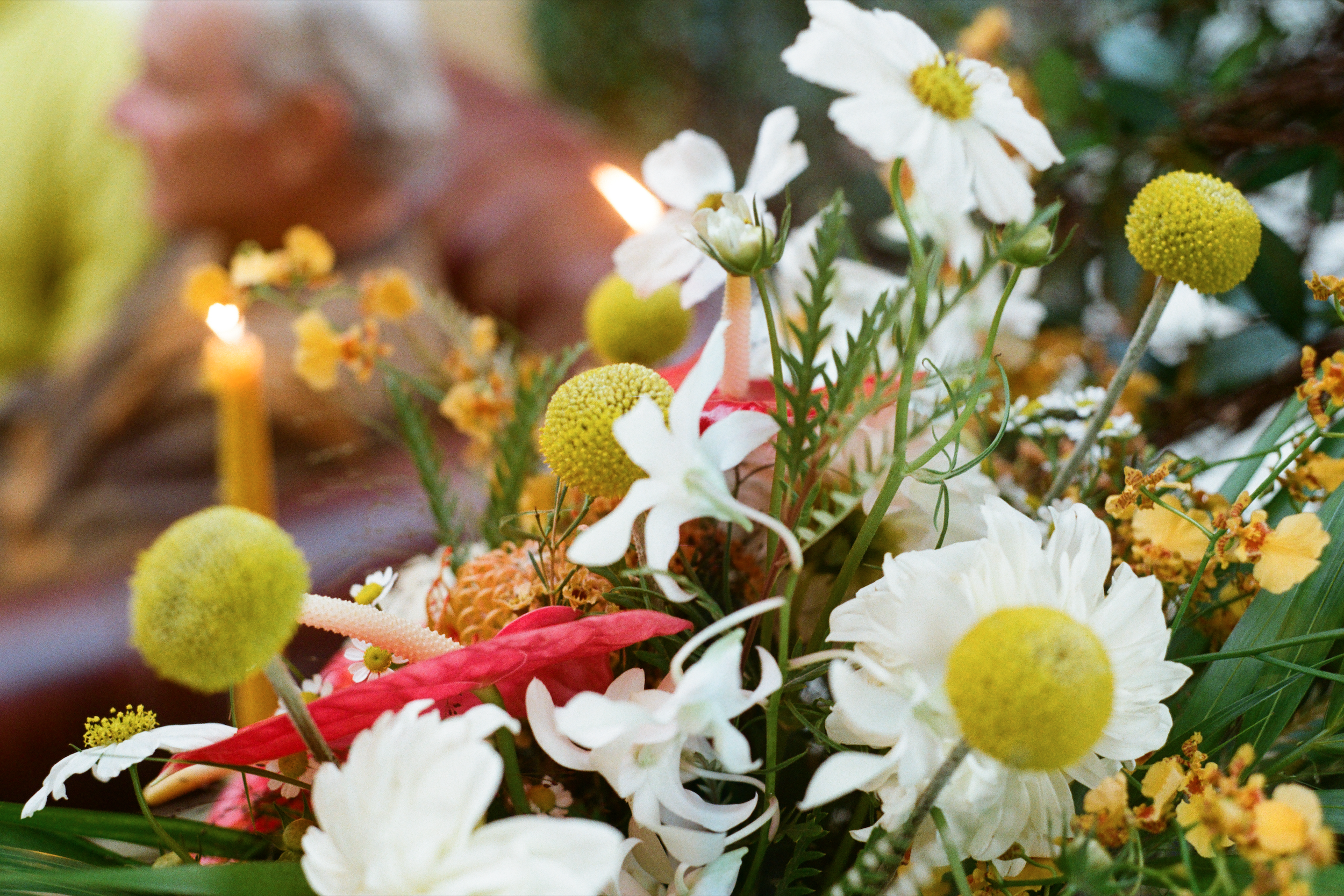 Close-up of a floral arrangement with white, yellow, and pink flowers, including daisies and other blossoms, with blurred background and small candles.