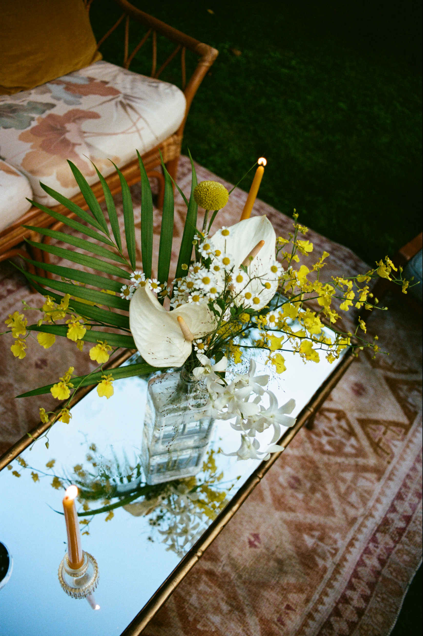 A floral arrangement in a glass vase on a mirrored table with candles, surrounded by a patterned rug, a wicker sofa, and green grass outside.