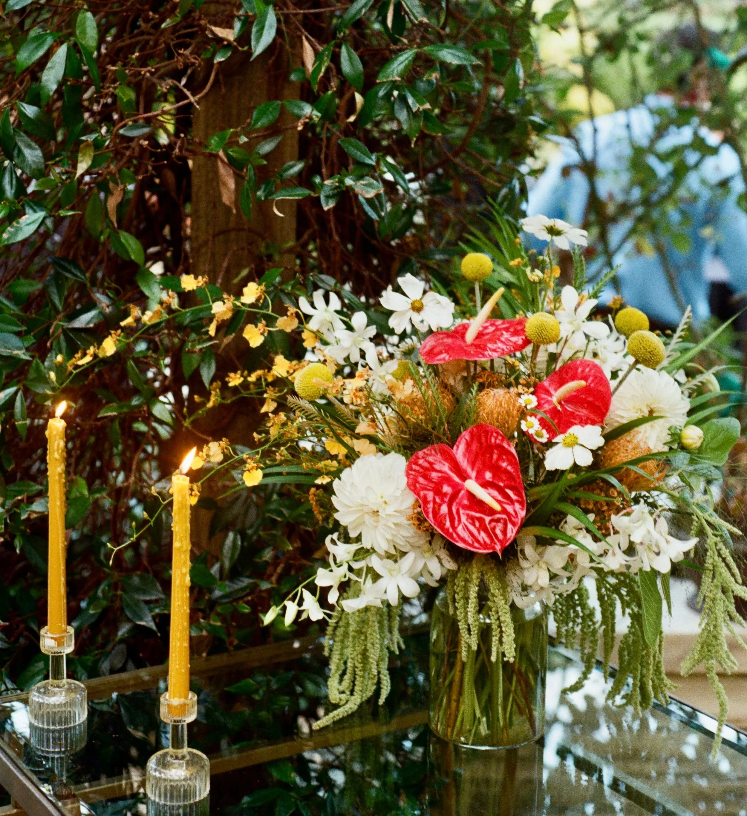 A colorful floral arrangement in a glass vase with white, red, yellow, and green flowers and greenery, set on a glass table with two lit yellow candles in glass holders nearby, against a background of leafy green plants.