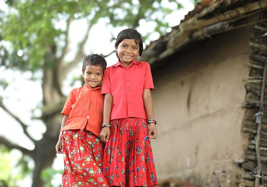 Two smiling girls stand outside of a rural home in South Asia