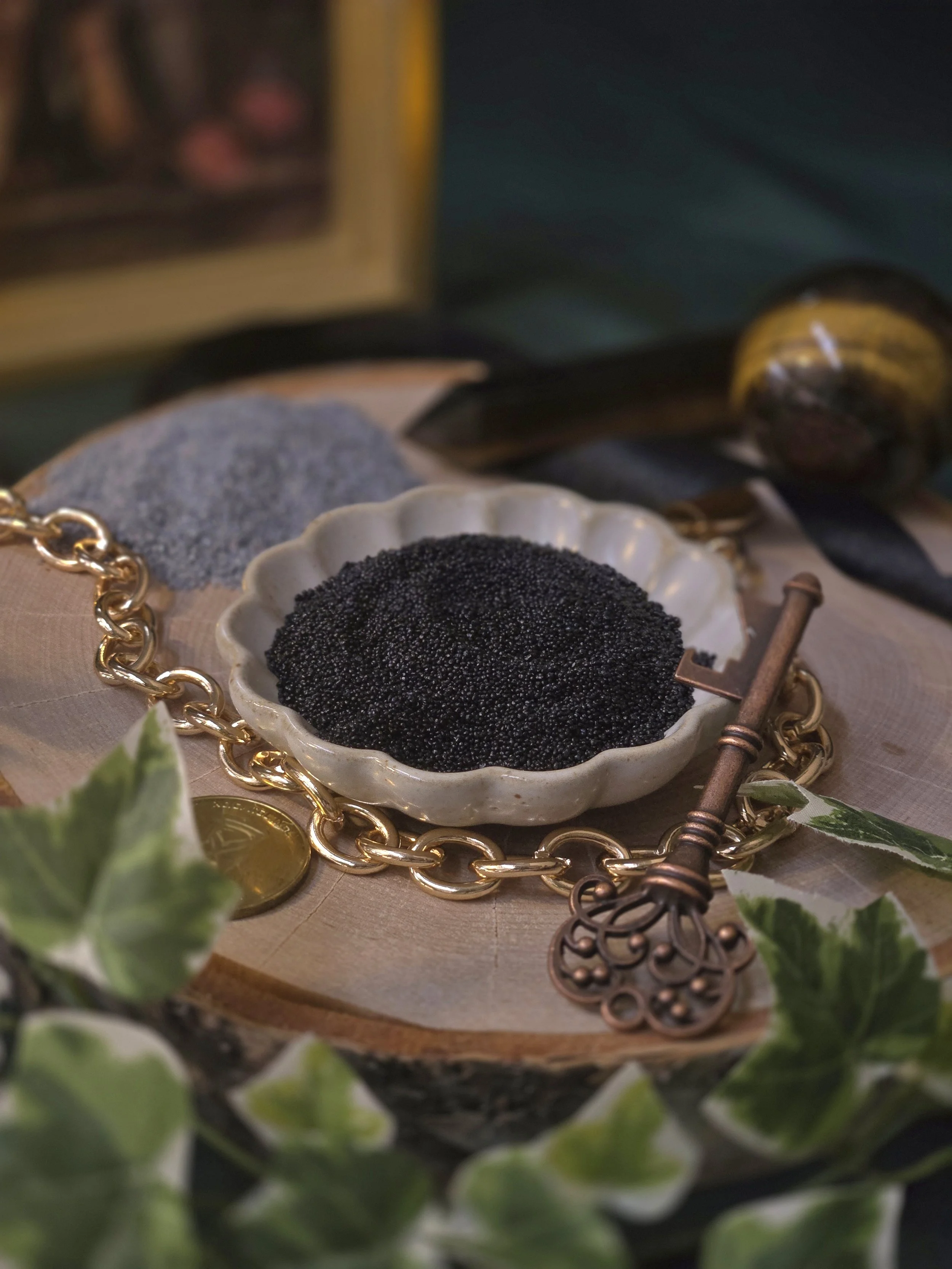 An antique white dish holds a pool of black pearled wax, with a neat pile of charcoal wax beside it on a birch round, styled with tiger eye crystals, an antique brass key, and a gold chain—evoking moody, dark academia vibes for magical events.