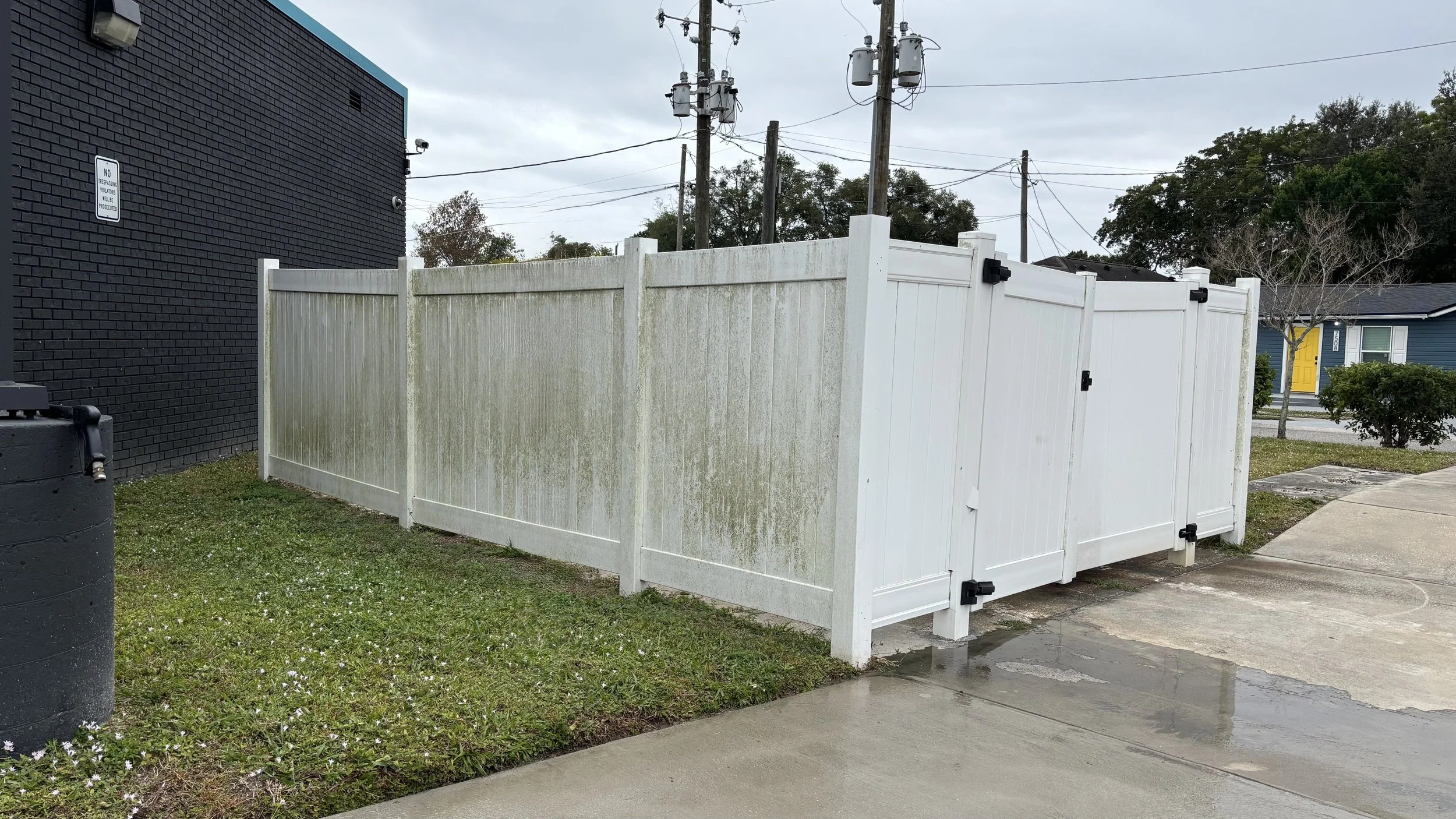 White vinyl fence with a gate surrounding a small grass area next to a black brick building. A large black rain barrel is in the foreground, and a