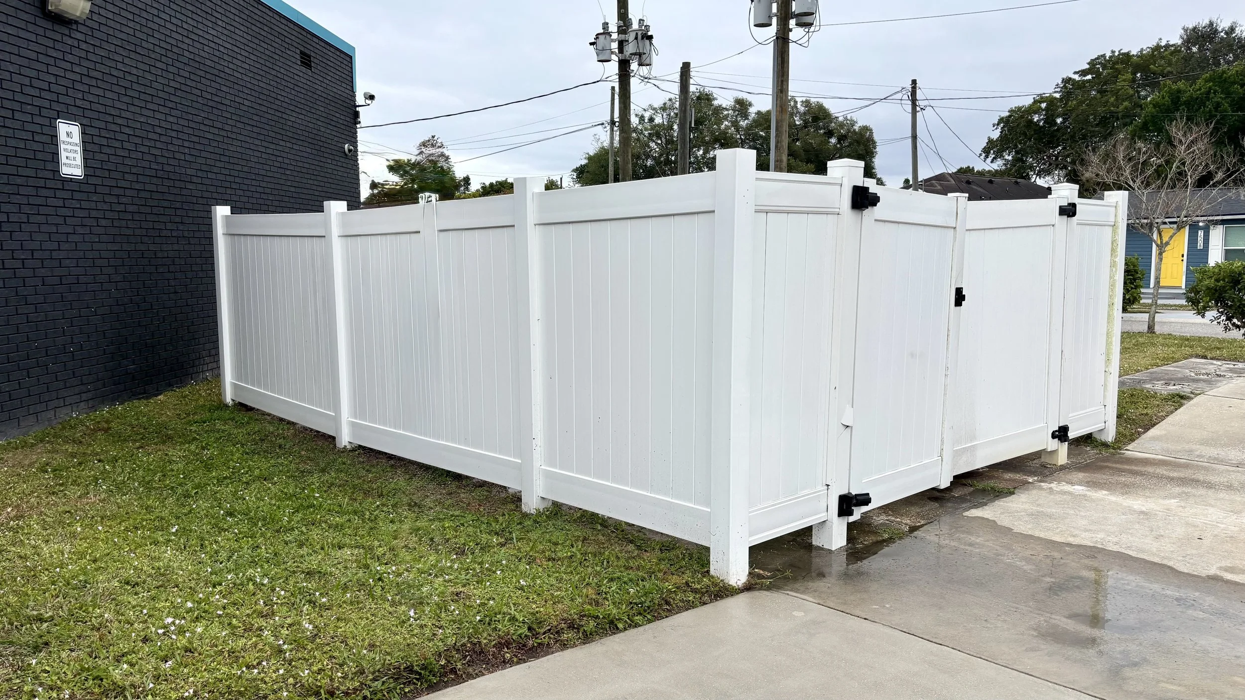 White vinyl privacy fence around a corner of a property, with a sidewalk and grassy area in front, and a brick building on the left side.