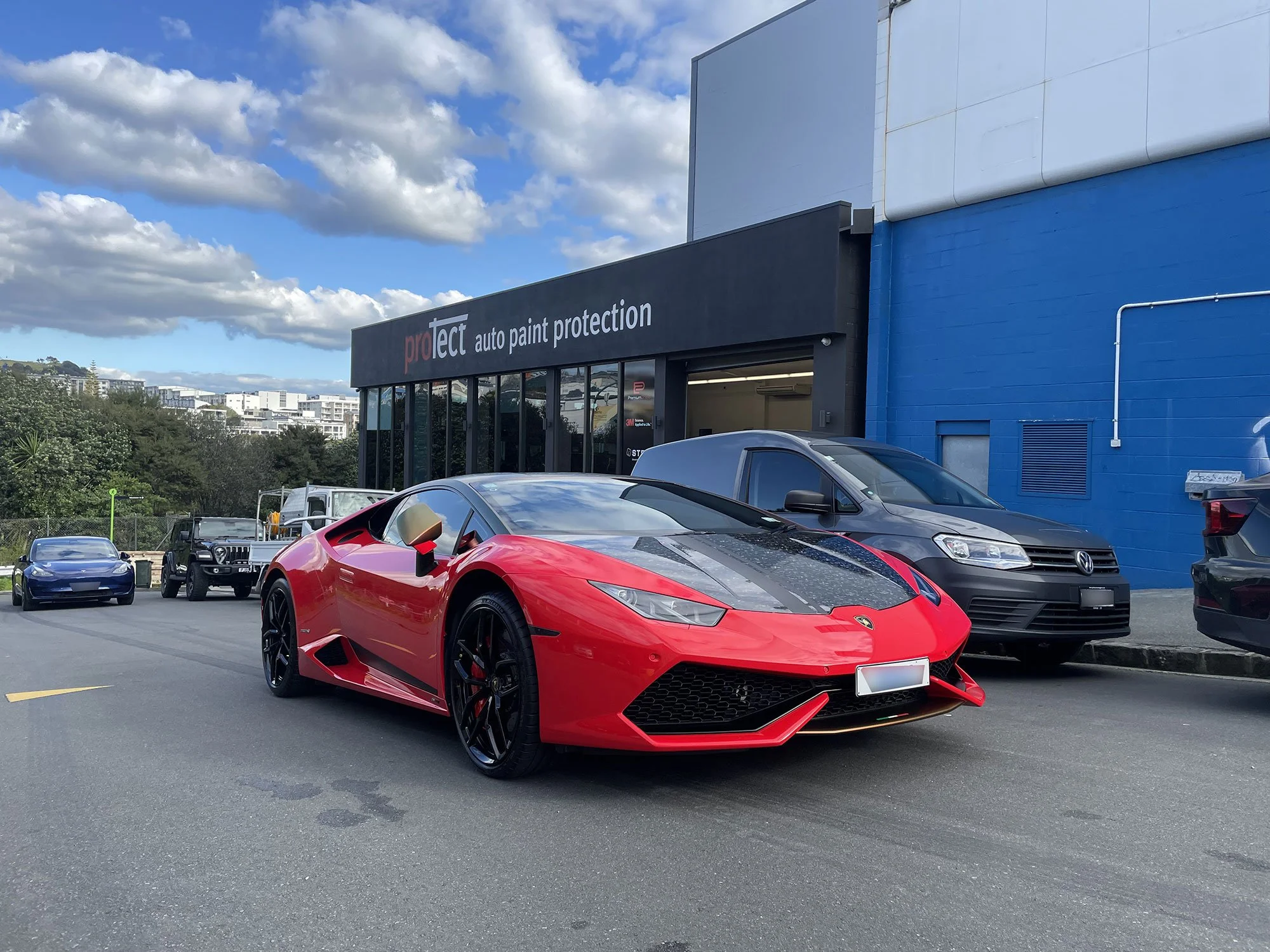 A red Lamborghini sports car parked on the street in front of an auto paint protection shop with black and blue exterior walls, alongside a gray van and other vehicles.
