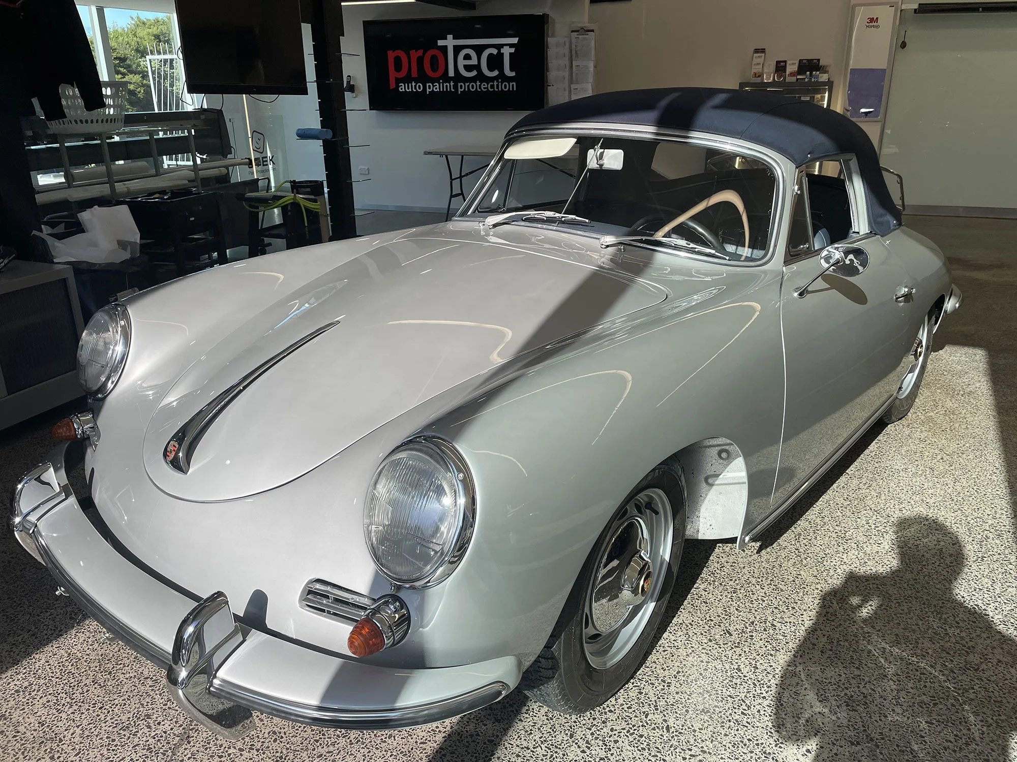 Silver vintage convertible car with black soft top parked indoors, with reflections of ceiling lights on its smooth surface.