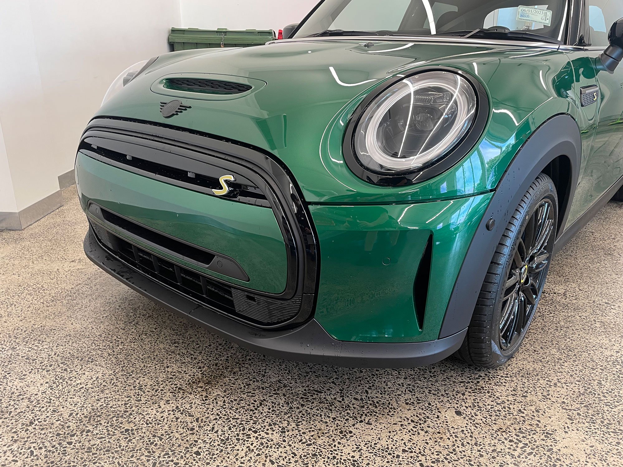Green Mini Cooper S parked indoors, front view showing the headlights, grille, and black wheel with tire.