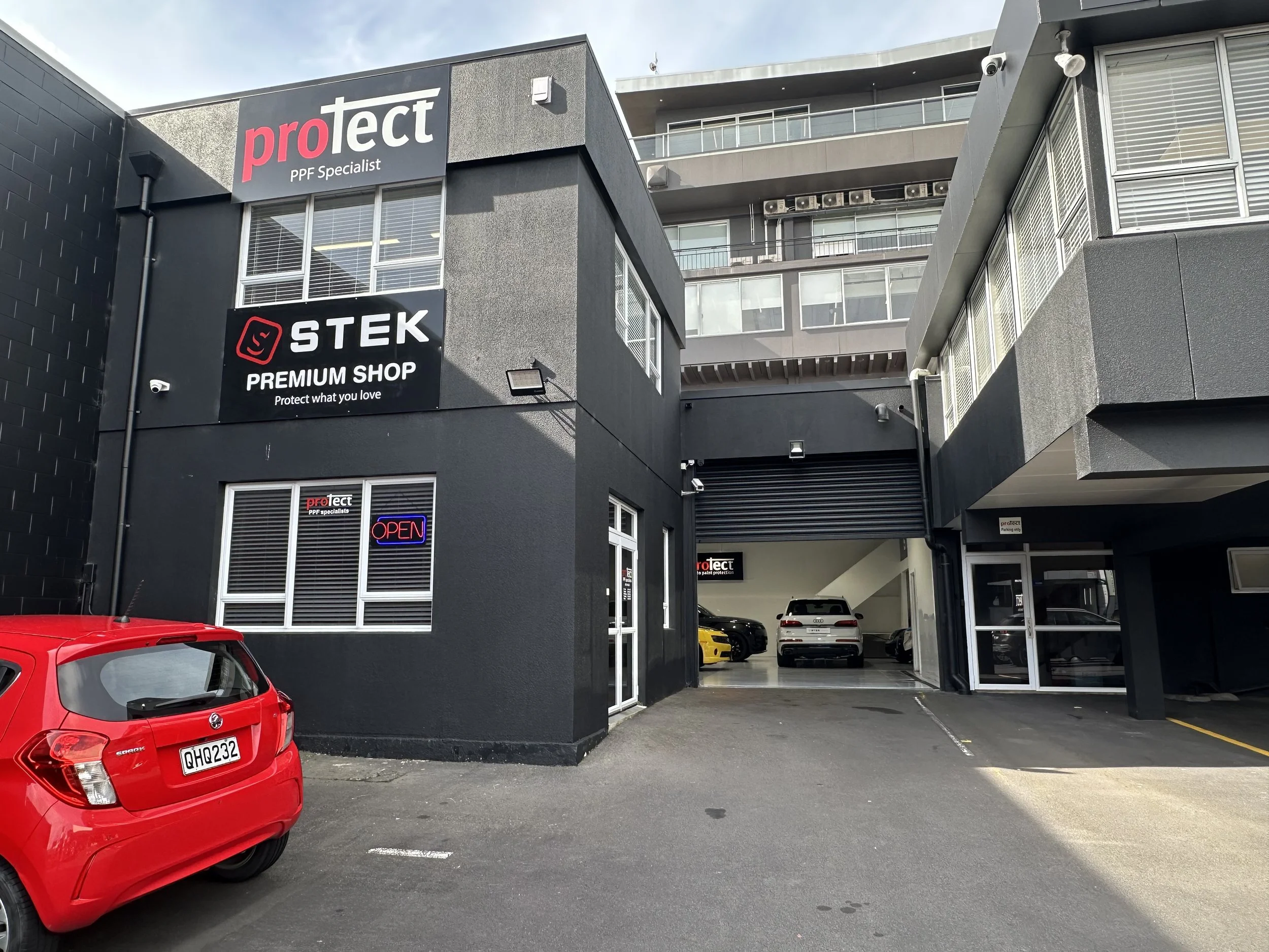 Exterior view of a black commercial building with signs for Protect PPF Specialist, STEK Premium Shop, and an illuminated 'Open' sign, with a red car parked in the foreground and other cars visible inside a garage area.