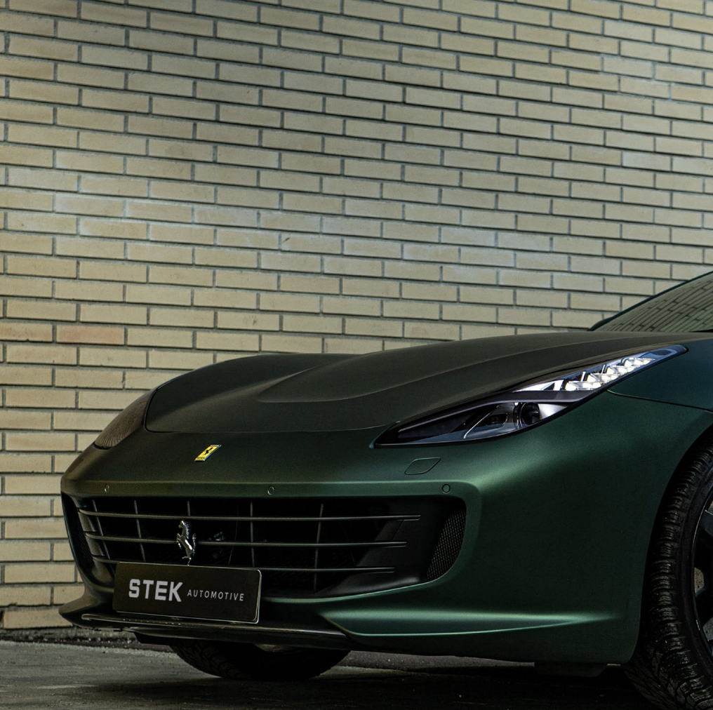 Front left side of a green Ferrari sports car parked against a brick wall, displaying sleek headlights and grille.