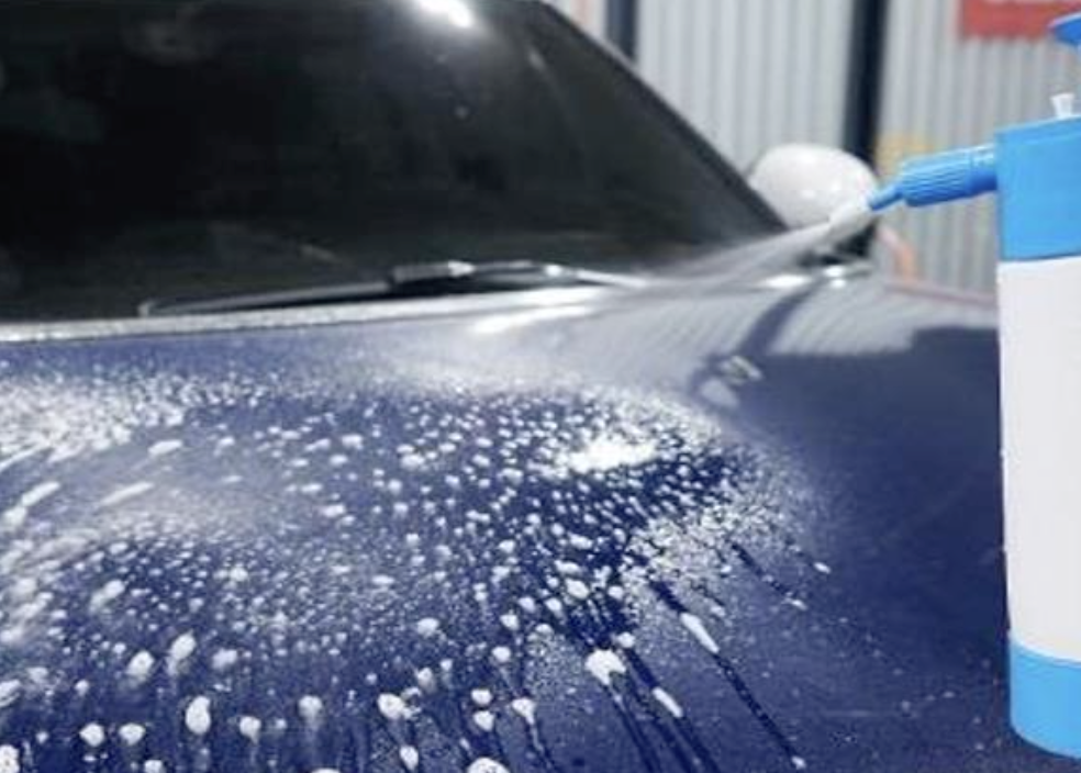 A person spraying a cleaning solution with a blue spray bottle onto the hood of a dark-colored car.