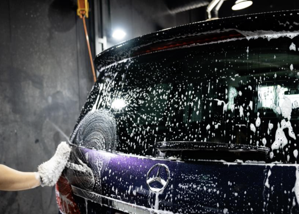 Person washing a black Mercedes-Benz car with soap and a sponge in a garage.