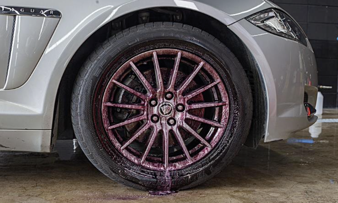 A close-up of a sports car's rear wheel with pink water or soap foam on the rim and tire, showing the car's white body and part of the headlight.